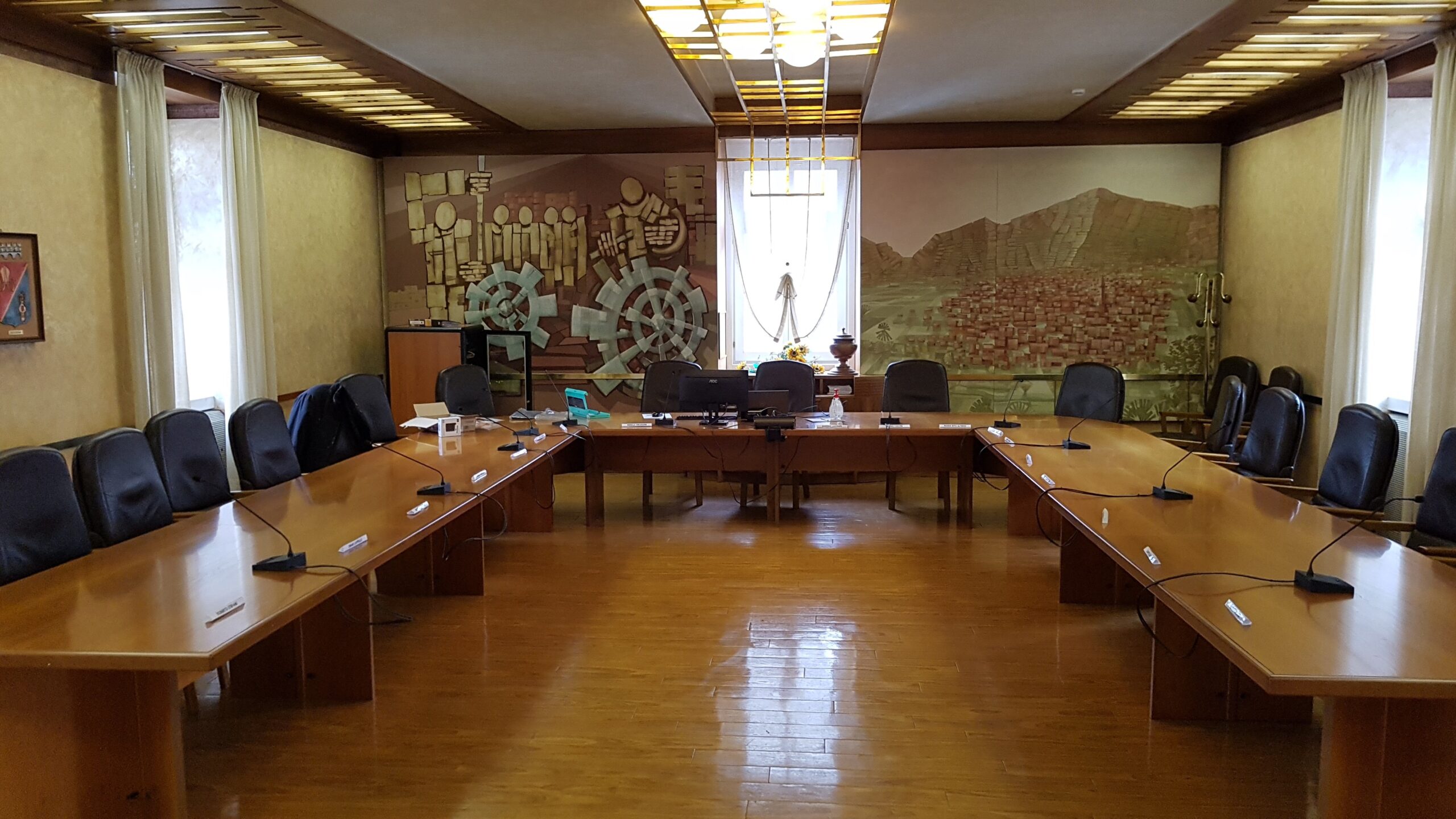 Vista frontale della sala consigliare di Tione con murale decorativo e microfoni da tavolo