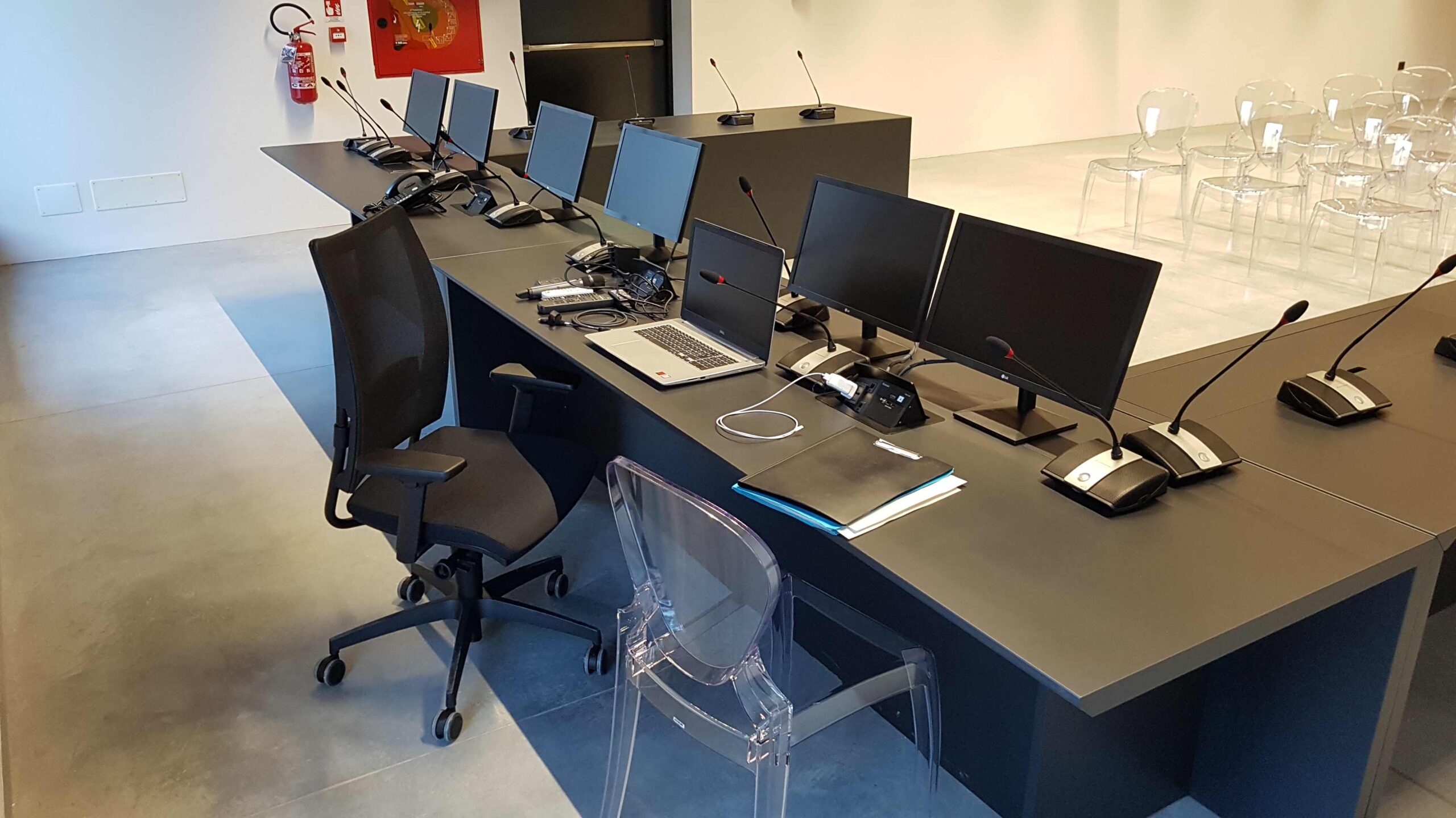 Dettaglio postazioni relatori con monitor, microfoni e sedute nella sala consiliare di Torbole